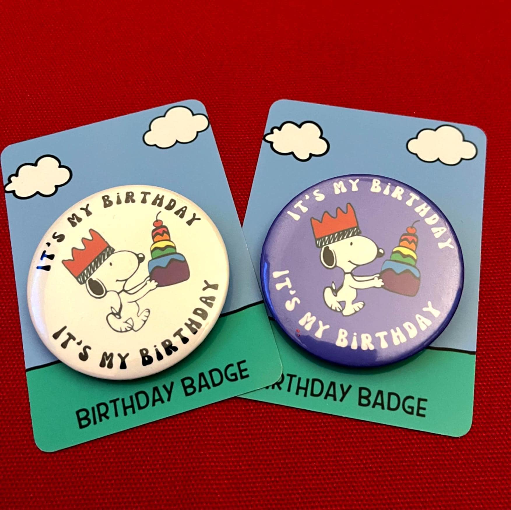 Two birthday badges with Snoopy holding a birthday cake on blue cards with clouds, against a red background.